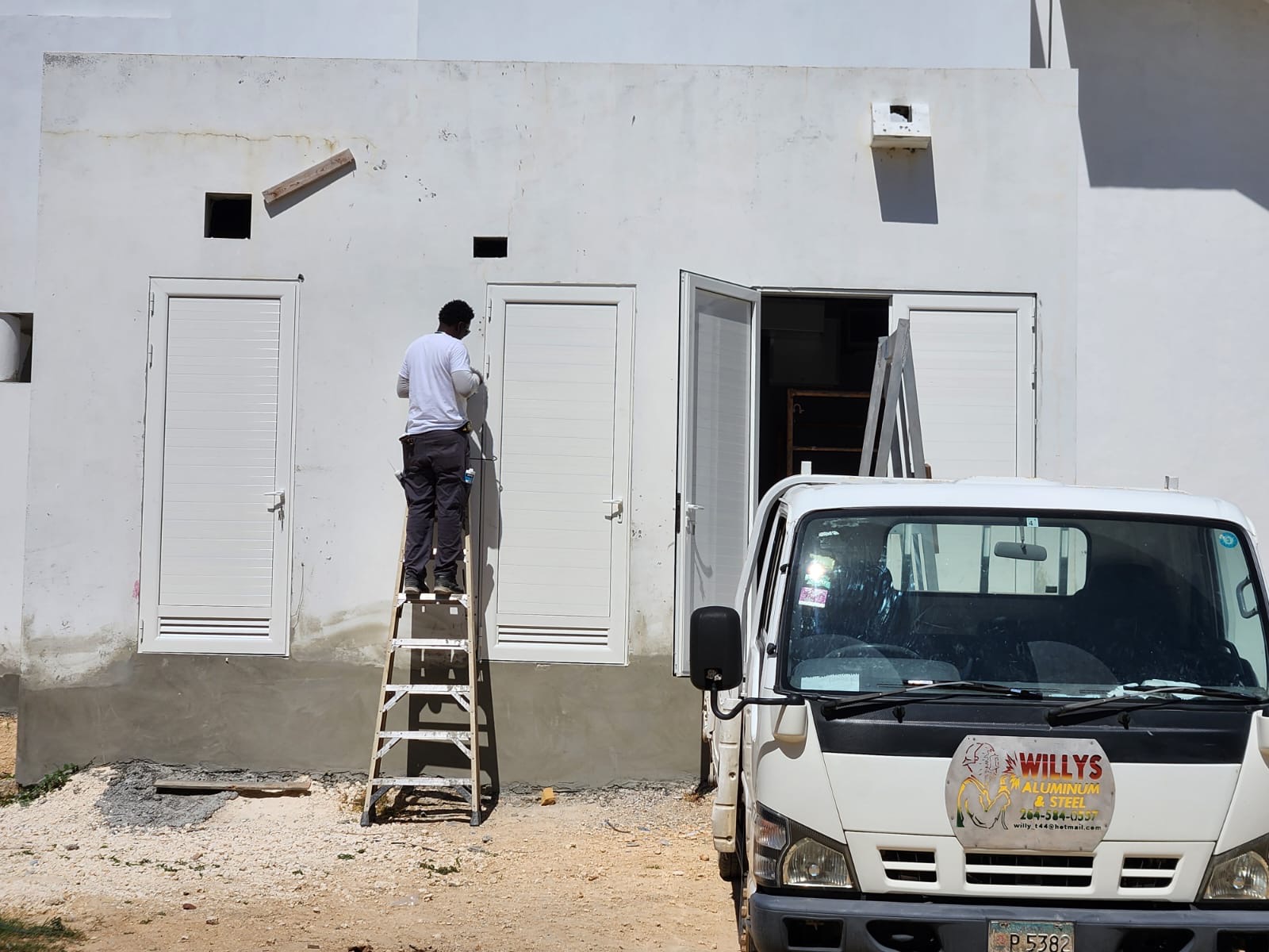 A Willy's Aluminium and Steel fitter installing white aluminium louvre doors on site in Anguilla, with the company truck parked alongside