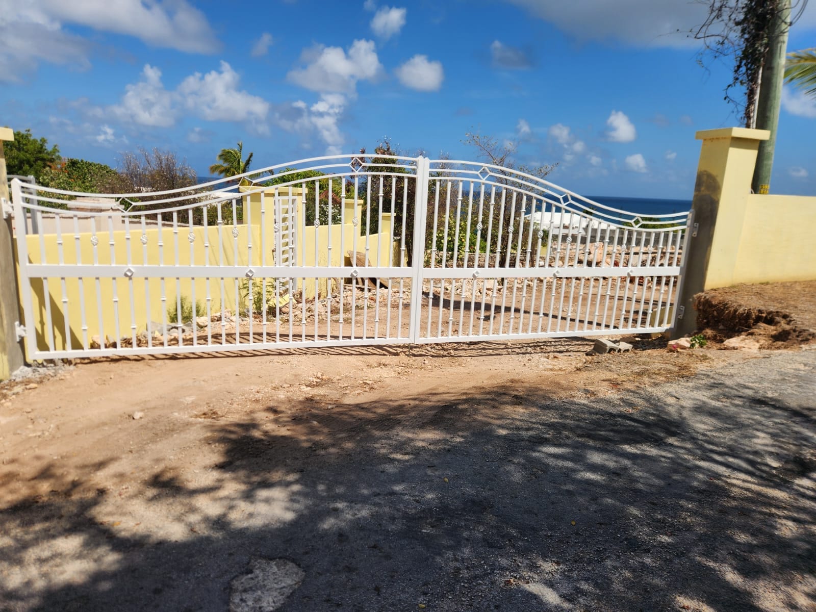 White aluminium double driveway gate with decorative scrollwork beside a yellow wall in Anguilla